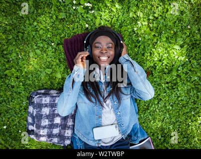 Vista dall'alto di sorridere donna africana giacente su erba verde per ascoltare musica con le cuffie. Foto Stock