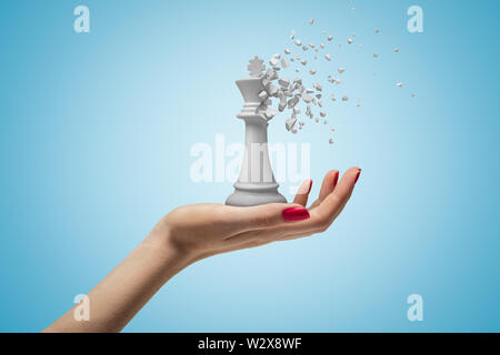 Primo piano della donna di mano rivolto verso l'alto e mantenendo grande bianco re di scacchi che ha iniziato a sciogliere in pezzi su fondo azzurro. Foto Stock