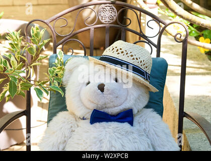 Orsacchiotto di peluche che indossa cappello per il sole sdraiato su una sedia in un giardino mostra come a rallentare, relax, sorriso con contentezza e calma Foto Stock