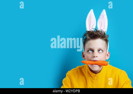 Un ragazzo in una giacca gialla con strabismo e Orecchie di lepre, tenendo una carota nella sua bocca su uno sfondo blu Foto Stock