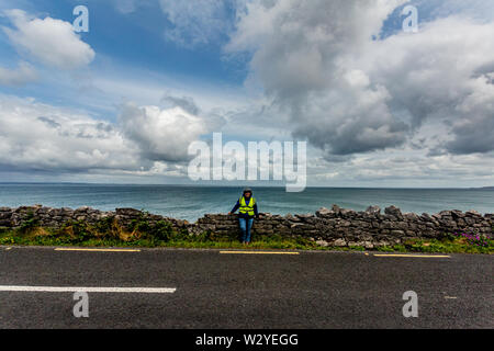 Ciclista femmina avente una pausa sulla fascia costiera R477 strada tra Bothar nA hAillite e Fanore, geosite e geoparco, Wild Atlantic modo, giornata di primavera Foto Stock