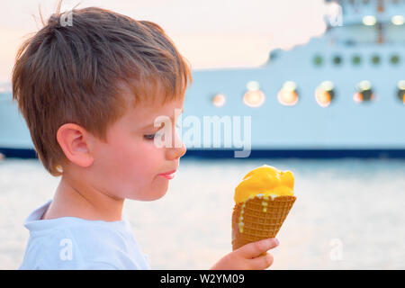 Ragazzo con gelato con yacht su sfondo. Sweet Food sfondo. Bambino mangiare gelato sulla riva del mare con ferry boat sullo sfondo. Dolce Vita Foto Stock