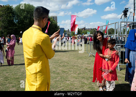 Londra. Tower Hamlets. I campi di tessitori. Boishakhi Mela. Un uomo vestito in una smart shawar kameez prende una foto sul suo telefono cellulare della sua famiglia. Foto Stock