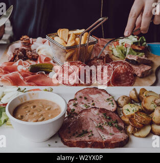 Close up dei maschi di mani tenendo una forcella, mangiare carne fredda piatto con chip, piastra con collare di maiale, patate e fagioli accanto, il fuoco selettivo. Foto Stock