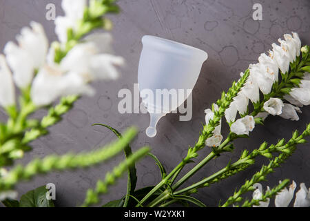 Bianco coppa mestruale per le donne accanto a fiori di colore bianco su sfondo grigio. Alternativa ecologica per le guarnizioni e i tamponi. Zero rifiuti Nozione Foto Stock