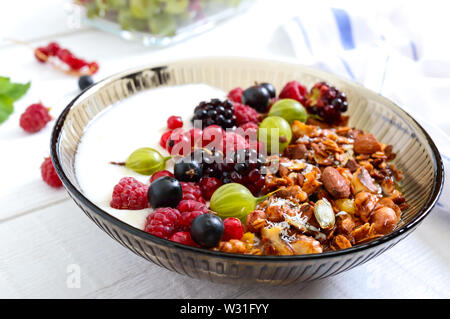 Yogurt, muesli, frutti di bosco freschi in un recipiente su un bianco sullo sfondo di legno. Delizioso e salutare prima colazione. Una corretta alimentazione. Menu dietetici. Foto Stock
