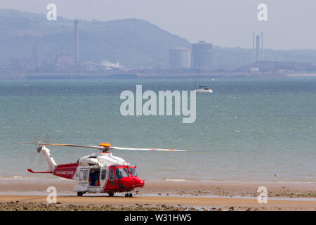 Sua Maestà i guardacoste Augusta Westland di colore rosso e bianco di atterraggio per elicotteri su una spiaggia con il mare e il complesso industriale di sfondo Foto Stock