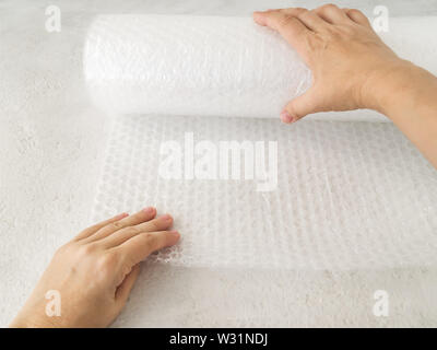 Donna mani di svolgere una bobina di bianco trasparente Bubble wrap su una ruvida sfondo bianco. Materiale per l'imballaggio di oggetti fragili per un trasporto sicuro. Foto Stock