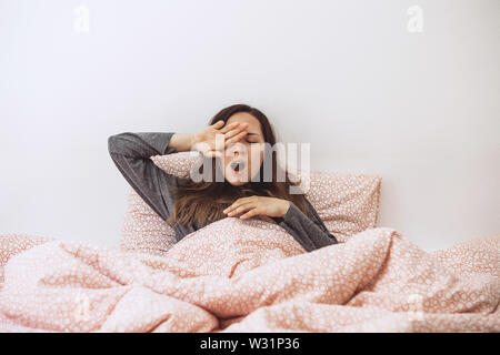 La ragazza è a letto e sbadigli. Ella si copre la bocca con la mano. La mattina presto e lei è sonnolenta. Foto Stock