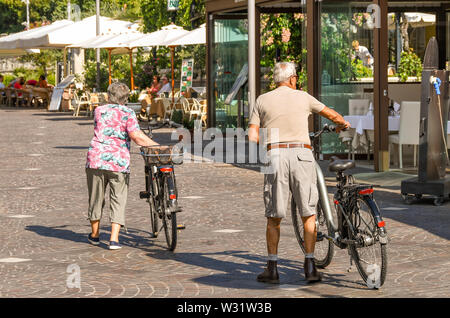 Il LAGO DI GARDA, Italia - Settembre 2018: uomo maturo e la donna wheeling biciclette passato ristoranti sul lago di Garda sul Lago di Garda. Foto Stock