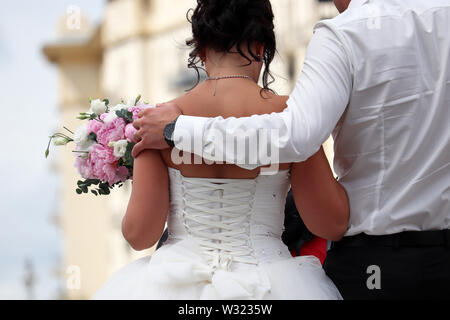 Appena una coppia sposata, sposo abbraccia la vita della sposa in piedi con bouquet di fiori, in vista posteriore Nozze in una città, il concetto della nuova famiglia Foto Stock