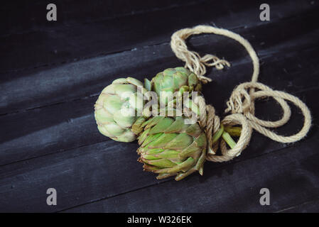 Close up fresco verde carciofi con corda intrecciata sul legno nero tavolo da cucina alleggerita dalla mattina la luce della finestra. Agricoltore raccolto, il mercato locale. S Foto Stock