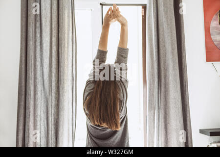 La ragazza si è svegliato e sta alla finestra e ha sollevato le sue mani verso l'alto. Foto Stock