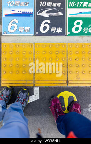 Utsunomiya, Giappone - Aprile 4, 2019: Guardando verso il basso ad alto angolo di visione del giovane, persone gambe o piedini in piedi sulla piattaforma con la stazione ferroviaria di numeri di auto da Foto Stock