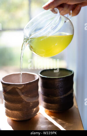 Primo piano delle mani femminili holding teiera di vetro versando il giapponese Sencha tè verde in tazze o teacups a casa per la prima colazione la mattina dalla finestra Foto Stock