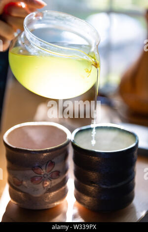 Primo piano della persona mani teiera di vetro versando il giapponese Sencha tè verde in tazze o teacups a casa per la prima colazione la mattina dalla finestra Foto Stock