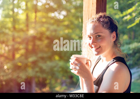 Donna in piedi sul portico di casa in cottage di legno mattina bere caffè o tè con luce dell'alba Foto Stock