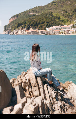 La ragazza si siede su un litorale roccioso e ammira una vista bellissima della città costiera di Petrovac in Montenegro. Foto Stock
