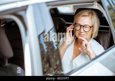Senior business Donna che parla con il telefono cellulare mentre si è seduti sul sedile posteriore di una macchina moderna. Concetto di vita aziendale in pensione Foto Stock