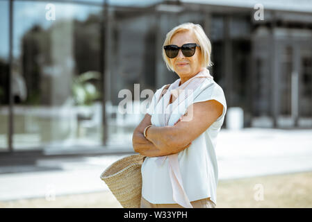 Ritratto di un felice e senior eleganti donna vestita in camicia bianca con occhiali da sole e la sacca all'aperto Foto Stock
