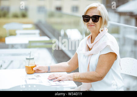 Ritratto di un elegante senior donna vestita di bianco seduto con drink estivo sulla terrazza all'aperto Foto Stock