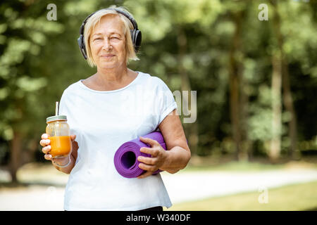 Ritratto di un attivo senior donna in abbigliamento sportivo in piedi con materassino yoga e succo di frutta all'esterno. Concetto di uno stile di vita sano in pensione Foto Stock