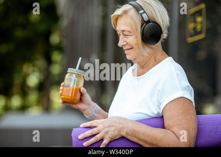 Ritratto di un attivo senior donna in abbigliamento sportivo in piedi con materassino yoga e succo di frutta all'esterno. Concetto di uno stile di vita sano in pensione Foto Stock