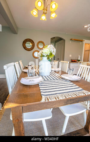 La sala da pranzo interna di una casa marrone con tavolo in legno e sedie bianche. Un lampadario a bracci è montato sul soffitto sopra il tavolo runner, fiori, vetro Foto Stock
