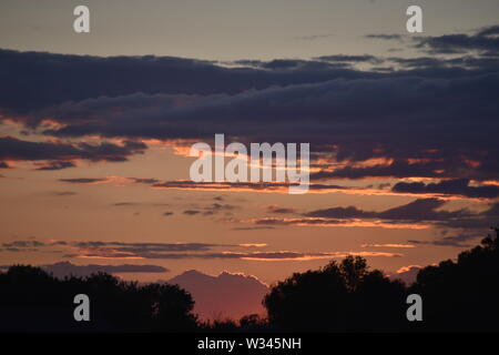 Tramonto sul flat suffolk campi con alcuni alberi in lontananza, viola scuro nuvole e un bello e caldo bagliore arancione nel cielo. Foto Stock