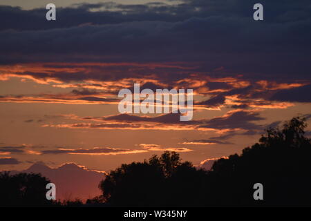 Tramonto sul flat suffolk campi con alcuni alberi in lontananza, viola scuro nuvole e un bello e caldo bagliore arancione nel cielo. Foto Stock