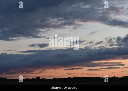 Tramonto sul flat suffolk campi con alcuni alberi in lontananza, viola scuro nuvole e un bello e caldo bagliore arancione nel cielo. Foto Stock