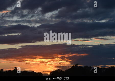 Tramonto sul flat suffolk campi con alcuni alberi in lontananza, viola scuro nuvole e un bello e caldo bagliore arancione nel cielo. Foto Stock