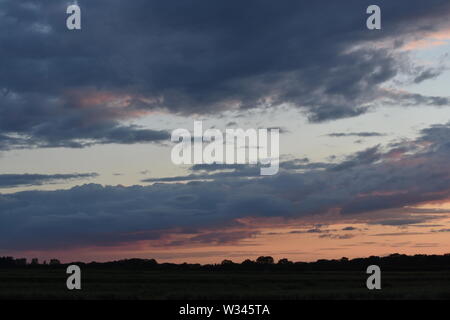 Tramonto sul flat suffolk campi con alcuni alberi in lontananza, viola scuro nuvole e un bello e caldo bagliore arancione nel cielo. Foto Stock