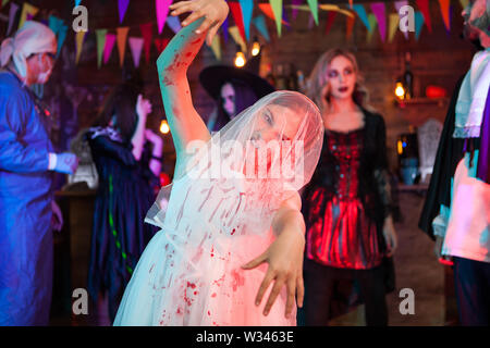 Scary creepy bambina abiti da sposa con Halloween trucco in operazioni di diserbatura abito. Crazy dottore in background. Foto Stock