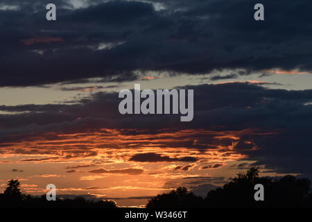 Tramonto sul flat suffolk campi con alcuni alberi in lontananza, viola scuro nuvole e un bello e caldo bagliore arancione nel cielo. Foto Stock