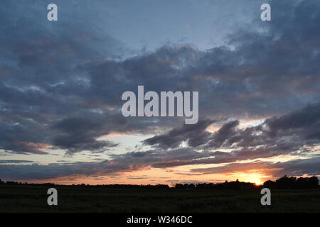 Tramonto sul flat suffolk campi con alcuni alberi in lontananza, viola scuro nuvole e un bello e caldo bagliore arancione nel cielo. Foto Stock