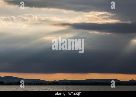 Sunray a vicino al tramonto con nuvole scure sullo sfondo di un cielo arancione e il lago Trasimeno (Umbria, Italia) al di sotto di Foto Stock