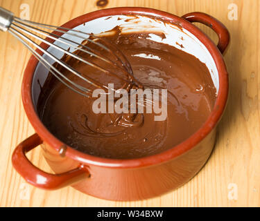 Pentola con frusta e cioccolato fuso su pannello di legno Foto Stock