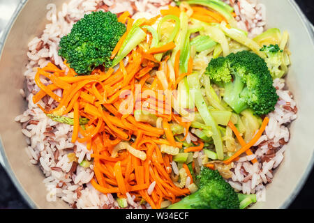Riso marrone con organici non mescolate pelati verdure tagliate a fette per la cottura in ghisa pentola. Luminose broccoli, la carota e la zucchina su sfondo di cereali. V Foto Stock