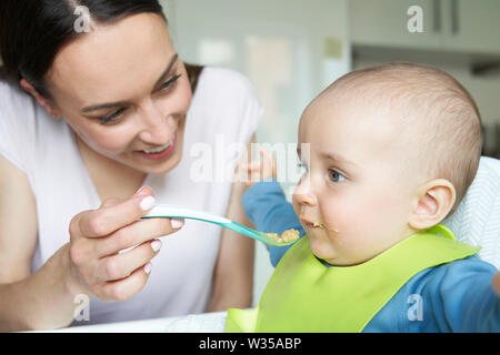 Sorridente 8 mese vecchio Baby Boy a casa in alta sedia essendo alimentato cibo solido da madre con cucchiaino Foto Stock