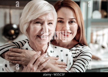 Bel sorriso. Calma Allegra donna di mezza età che abbraccia delicatamente il suo allegro anziana madre e sorridente Foto Stock