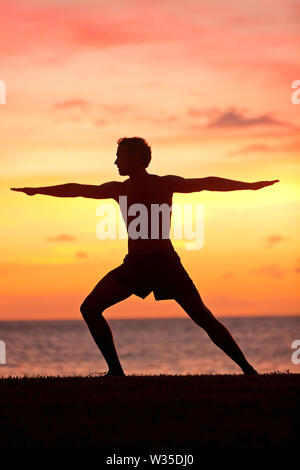 Uomo di Yoga formazione e meditando nella posizione del guerriero pongono al di fuori dalla spiaggia all'alba o al tramonto. Maschio di istruttore yoga che lavora fuori della formazione in oceano sereno paesaggio. Silhouette di modello da uomo contro sun. Foto Stock