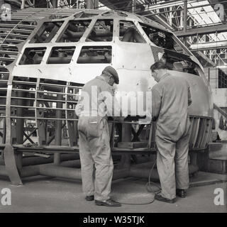 1950, storica aviazione del dopoguerra, due meccanici maschi in tuta, che lavorano all'esterno di una cabina di pilotaggio all'interno di una fabbrica aerospaziale o di un gancio, Inghilterra, Regno Unito. Foto Stock