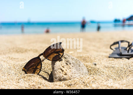 Occhiali da sole marrone tendeva verso una pietra su una spiaggia con le infradito e persone sfocate in background Foto Stock