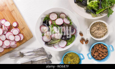 Mix di erbe con insalata fresca di ravanello organico, lattughe e spinaci, alcuni condimento, mandorle e olio vegetale. Close up su sfondo bianco, Donna mani Foto Stock