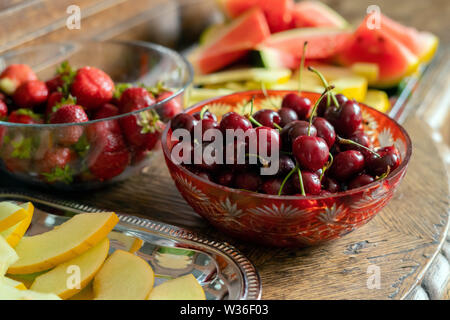 Freschi frutti estivi: ciliegie e fragole organico, fette di melone, anguria annata nella ciotola di cristallo sul vecchio tavolo in legno Foto Stock