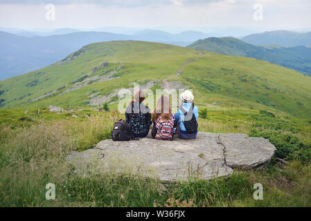Primo piano di tre giovani donne, femmina gli escursionisti con zaini in un cappello seduti insieme e guardando a pittoresche colline su di un bordo di una roccia. Nuvoloso rai Foto Stock