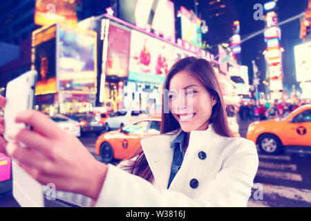 Donna felice turistica prendendo foto selfie con la compressa nella città di New York Manhattan, Times Square. Multietnica caucasico asiatica donna nella sua 20s gioiosi e sorridenti indossando la molla pea coat. Foto Stock