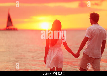 Luna di Miele romantica in amore tenendo le mani al tramonto sulla spiaggia. Sposa felice coppia giovane godendo del tramonto sull'oceano durante i viaggi vacanze vacanza. Interracial giovane. Foto Stock