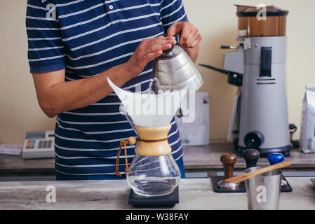 Chiudere l immagine del giovane maschio barista versando acqua bollente dal bollitore a gocciolare caffettiera sul tavolo di legno Foto Stock